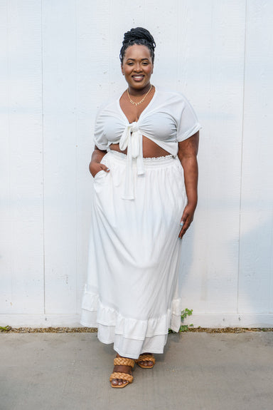 Plus size woman with black hair, wearing tied up top in white, right hand in pocket of maxi skirt in white, with legs crossed.